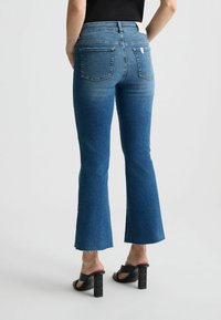 Blaue, ausgestellte Denim-Jeans mit hoher Taille, im verwaschenen Finish, zwei Gesäßtaschen und rohem Saum. Kombiniert mit schwarzen, klobigen Sandalen mit Absatz.