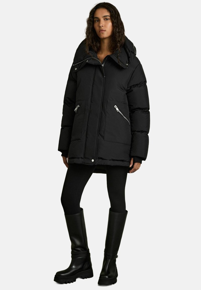 Svart puffjacka med hög krage, två framfickor med blixtlås och knappstängning. Kombinerad med svarta leggings och knähöga stövlar.