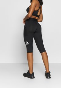 Kobieta w czarnych legginsach Adidas, czarnym staniku sportowym i czarnych butach biegowych, stoi z założonymi rękami, odwrócona plecami do aparatu.