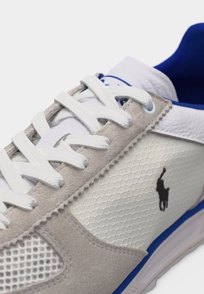Λευκό και γκρι αθλητικό παπούτσι με λευκά κορδόνια, μπλε εσωτερικό, υφασμένη πλαϊνή επιφάνεια και μαύρο λογότυπο παίκτη πόλο στο πλάι.