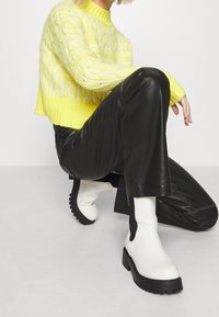 Person som knäböjer iklädd en gul kabelstickad tröja, svarta läderbyxor och vita klumpiga ankelskor mot en neutral bakgrund.