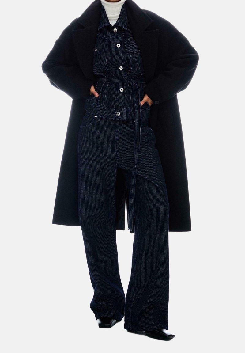 Personne portant un pantalon large noir, une veste assortie ceinturée avec des boutons blancs, un long manteau noir et un col roulé blanc, debout les mains dans les poches.