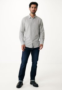 Camicia grigia chiaro con sottili righe bianche, abbinata a jeans blu scuro e stivali neri. La camicia ha colletto e maniche lunghe.