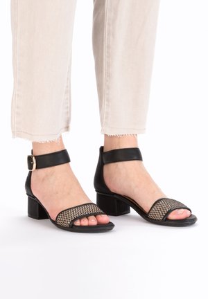 Schwarze, geflochtene Sandaletten mit offenem Zehenbereich und einem Blockabsatz, einem Knöchelriemen und einem strukturierten Muster auf der Vorderseite. Legere beige Cropped-Hosen.