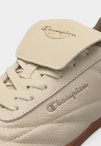 Μπεζ δερμάτινο sneaker Champion που δείχνει τα κορδόνια, το κεντημένο λογότυπο στο πλάι και το δερμάτινο καπάκι της γλώσσας με ανάγλυφη σήμανση Champion.
