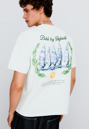 Junger Mann trägt weißes T-Shirt mit Segelboot-Grafik, grünem Lorbeerkranz, Sonnenillustration und dem Text "Bold by Default" auf dem Rücken.