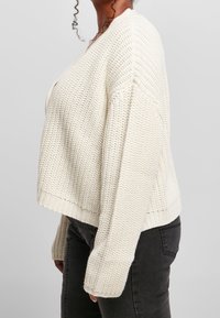 Kvinna som bär en kraftig off-white stickad cardigan med långa ärmar och svarta jeans, sedd från sidan mot en enfärgad bakgrund.