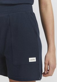 Middengrijze shorts van gestructureerde stof met een ribbelpatroon, voorzien van zijzakken en een zichtbaar label met de tekst "OXMO."