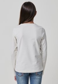 Person mit langen braunen Haaren, der ein einfaches weißes Langarmshirt und eine blaue Jeans trägt, steht mit dem Rücken zu einem einfarbigen Hintergrund.