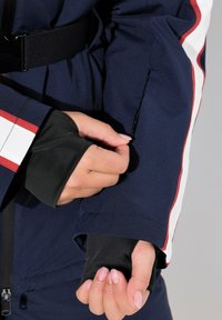 Námořnická modrá sportovní bunda má červené a bílé pruhy, elastické černé manžety a kapsu na zip. Detailní záběr na ruku upravující rukáv.