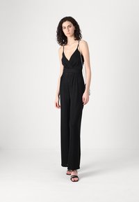 Crni kombinezon s preklopnim prednjim dijelom, uskim naramenicama, širokim nogavicama i teksturiranim materijalom; uparen s crnim sandalama s trakicama.