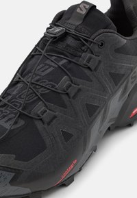 Chaussure de sport noire avec une tige texturée, entourée d'accents synthétiques noirs. Dispose d'un système de laçage rapide et d'une semelle extérieure en caoutchouc proéminente.