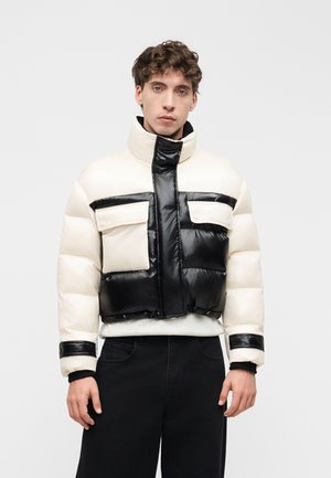 Joven con chaqueta acolchada negra y blanca con bolsillos aplicados, pantalones negros, de pie frente a un fondo claro y liso.