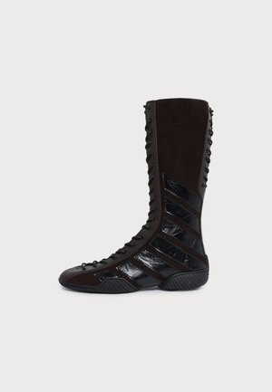 Une botte à lacets marron foncé montant jusqu’au genou, avec des empiècements diagonaux noirs brillants et une semelle noire rainurée, vue de profil.