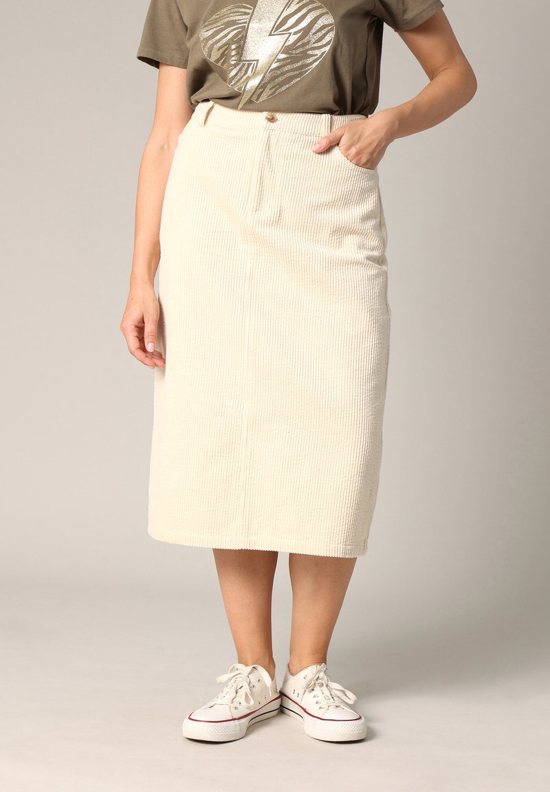 Jupe en velours côtelé crème, longueur genou, avec une texture à côtes verticales, dotée d'une ceinture, d'une poche et associée à des baskets blanches.