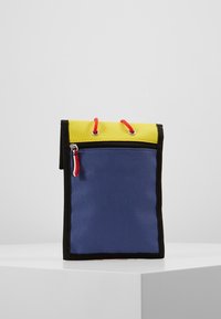 Borsa di stoffa colorata con parte superiore gialla, corpo blu, bordi neri e stringhe di chiusura rosse. Presenta una tasca con zip sulla parte anteriore.