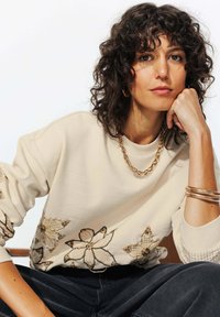 Sweatshirt beige avec broderie florale en or et teintes pastel, poignets côtelés et coupe décontractée, complété par des bijoux en or superposés.