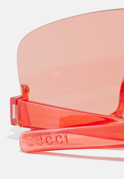 Gafas de sol tintadas en rojo con lentes de gran tamaño, montura de plástico y logotipo grabado en la patilla. Textura suave y detalles de hardware mínimos.