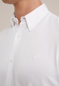 Camisa blanca de botón, hecha de tela suave, que presenta un cuello clásico y un sutil logo bordado en el pecho.