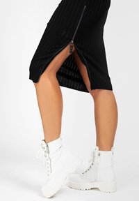 Jupe midi noire côtelée avec une fente haute, ornée d'un détail de fermeture éclair argentée. Associée à des bottines blanches à lacets avec des semelles épaisses.