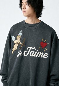Personne portant un sweat-shirt foncé avec un cupidon brodé, un cœur rouge et le texte "Je T'aime" sur la poitrine, regardant sur le côté devant un fond uni.