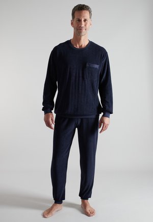 Man standing barefoot wearing a navy blue long-sleeve top with chest pocket and matching pants against a plain gray background.