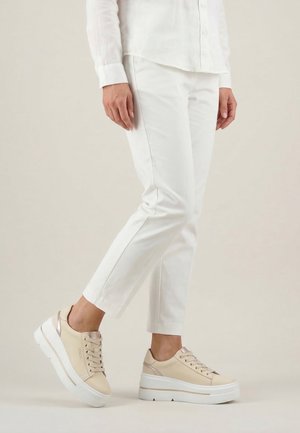 Osoba ubrana w białe krótkie spodnie i beżowe platformowe trampki z sznurowadłami stoi na neutralnym tle.