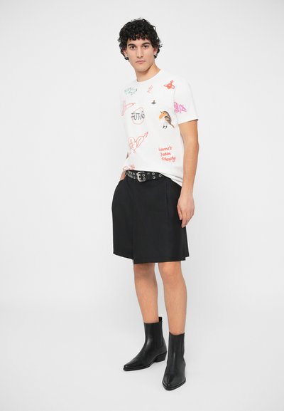 Hombre con camiseta gráfica blanca, pantalones cortos negros con cinturón y botas negras hasta el tobillo, de pie contra un fondo claro y liso.
