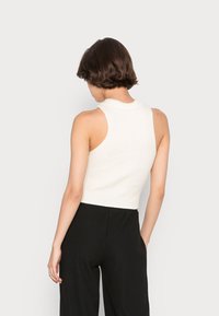 Hladký, bezrukávový bílý pletený top s vysokým krkem. Nosený s černými kalhotami s širokými nohavicemi, pohled zezadu.