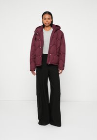 Giacca puffer bordeaux con cappuccio, caratterizzata da un design trapuntato e zip frontale, indossata sopra una maglietta grigia e pantaloni neri a gamba larga.