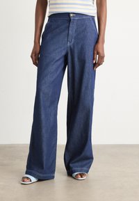 Weite dunkelblaue Jeans mit hoher Taille, ausgestattet mit Standardtaschen und sichtbaren Nähdetails. Dazu passende blaue Rutschsandalen.