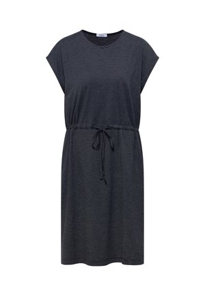 Lächelnde Frau in einem dunkelblauen, knielangen Kleid mit Kordelzug und beigen Slippern vor neutralem Hintergrund.