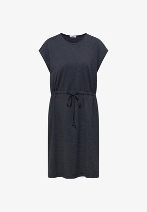 Femme souriante portant une robe bleu marine à cordon à hauteur des genoux et des chaussures beige sans lacets, debout devant un fond neutre.