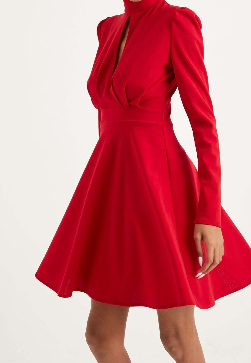 Femme portant une robe rouge vif à manches longues avec un col montant, une taille cintrée et une jupe évasée, debout devant un fond uni.