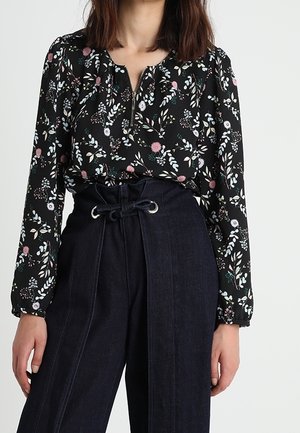 Frau mit einer langärmeligen schwarzen Bluse mit Blumenmuster und einer hochtaillierten dunklen Jeans mit Kordelzug, steht vor einem schlichten Hintergrund.