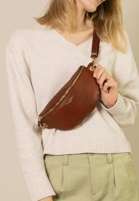 Marsupio in pelle marrone con texture in rilievo, zip dorata e tracolla regolabile, indossato a tracolla sopra un maglione leggero.