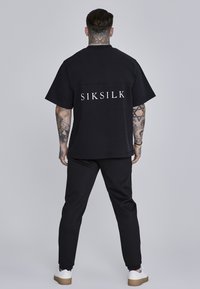 Črna majica s kratkimi rokavi z velikim logotipom "SIKSILK" na hrbtu, v kombinaciji s črnimi oprijetimi hlačami in belimi športnimi copati. Gladka tekstura blaga.