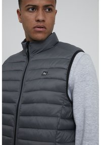 Chaleco acolchado gris con cuello alto y cierre de cremallera, que presenta un patrón de acolchado horizontal y un pequeño logotipo en el pecho.
