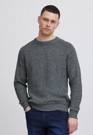 Strukturierter blau-grüner Strickpullover mit Rundhalsausschnitt und gerippten Bündchen, kombiniert mit dunklen Jeans. Hat eine lockere Passform und ein dezentes Muster.