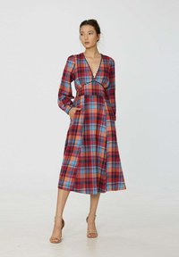 Robe à carreaux rouge, bleu et orange avec des manches longues, décolleté en V profond et poches. Tissu fluide, longueur genoux, corsage ajusté et jupe évasée.