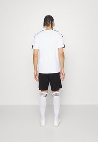 Maglietta sportiva bianca con accenti neri, abbinata a pantaloni shorts neri e calze bianche con strisce nere. Design di abbigliamento sportivo semplice e funzionale.