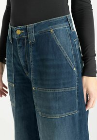 Pantalons cargo en denim bleu foncé avec des finitions dorées, dotés de multiples poches, d'une taille haute et d'une coupe décontractée.