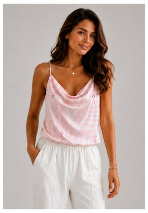 Femme aux longs cheveux foncés portant un haut sans manches rose à motifs et un pantalon blanc taille haute, debout devant un fond gris uni.