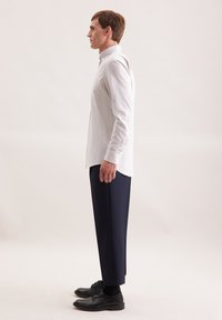 Camicia a quadri bianca con colletto, abbinata a pantaloni blu scuro e scarpe di pelle nere. Modello in posizione profilo su uno sfondo neutro.