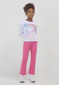 Junges Mädchen steht mit einem weißen Langarmshirt mit dem Aufdruck "Positive Vibes", pinken Hosen, weißen Socken und weißen Turnschuhen vor einem einfarbigen Hintergrund.