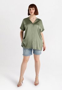 Camicia botton-up in satin verde oliva con scollatura a V e maniche arrotolate, abbinata a pantaloni shorts in denim e sandali con cinturini argentati.