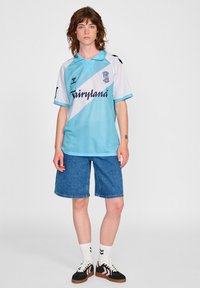 Ljusblå fotbollsdräkt med ett vitt diagonalt rand och marinblå bokstäver. Bärs med denimshorts och svarta sneakers med rosa accenter.