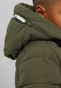 Nahaufnahme einer Person, die eine olivgrüne gepolsterte Jacke mit einem Reißverschluss und sichtbaren Nähten trägt.