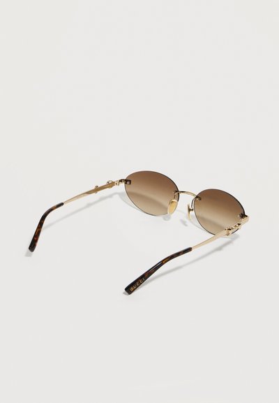 Στρογγυλοί φακοί με καφέ διαβάθμιση και χρυσό μεταλλικό σκελετό με άκρες από κέρατο χελώνας, με το λογότυπο Gucci στα πλαϊνά, γυαλιά ηλίου που ακουμπούν σε ανοιχτή επιφάνεια.