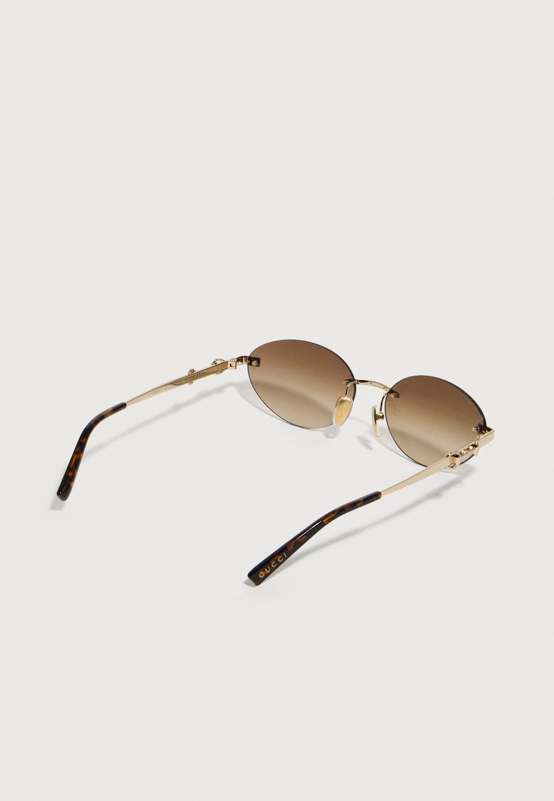 Verres ronds dégradés marron avec monture en métal doré et embouts en écaille de tortue, logo Gucci sur les branches, lunettes de soleil posées sur une surface claire.